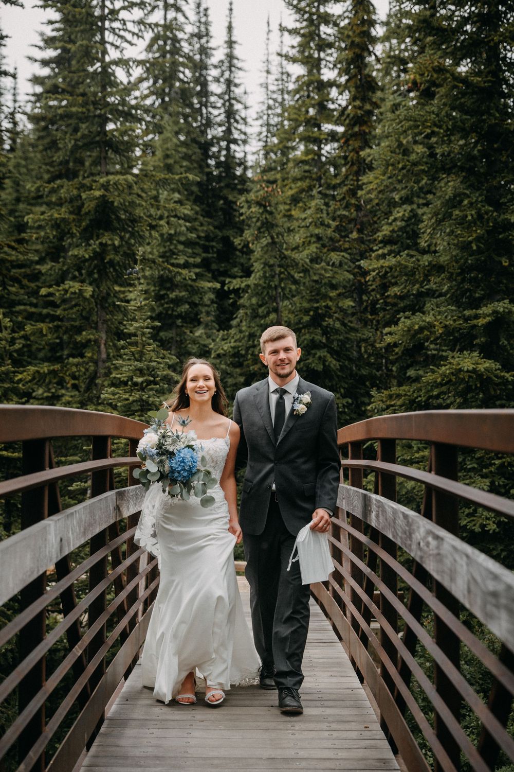 Newlyweds share playful moments on a wooden bridge surrounded by evergreen trees.