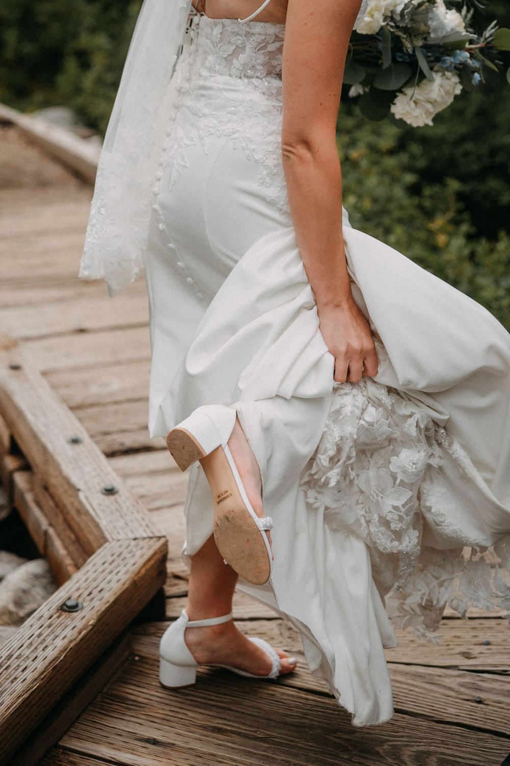 Close-up of a bride lifting her white wedding dress to show her shoes on wooden dock steps.