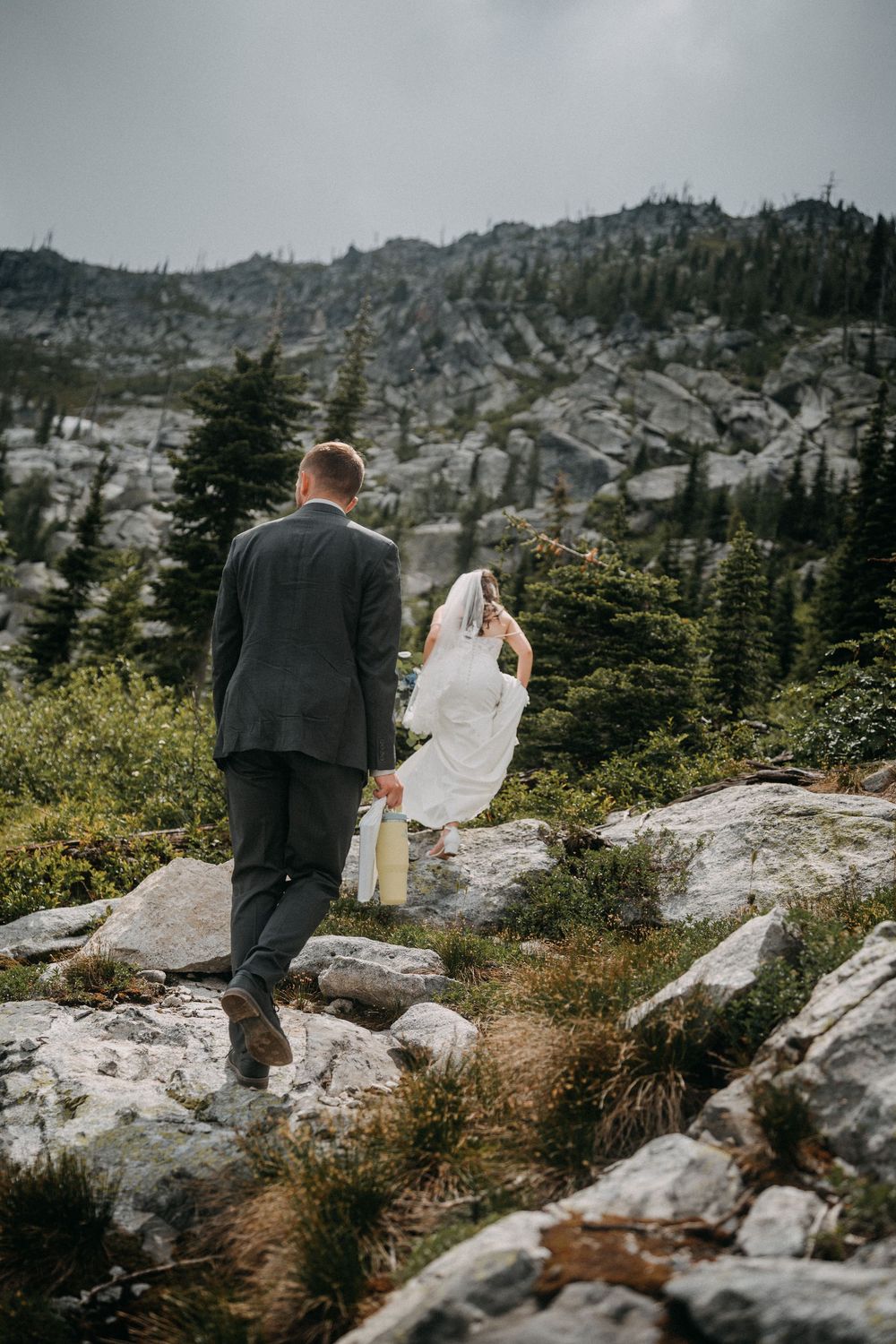 A couple walks along a rocky mountain trail surrounded by evergreen trees and rugged terrain.