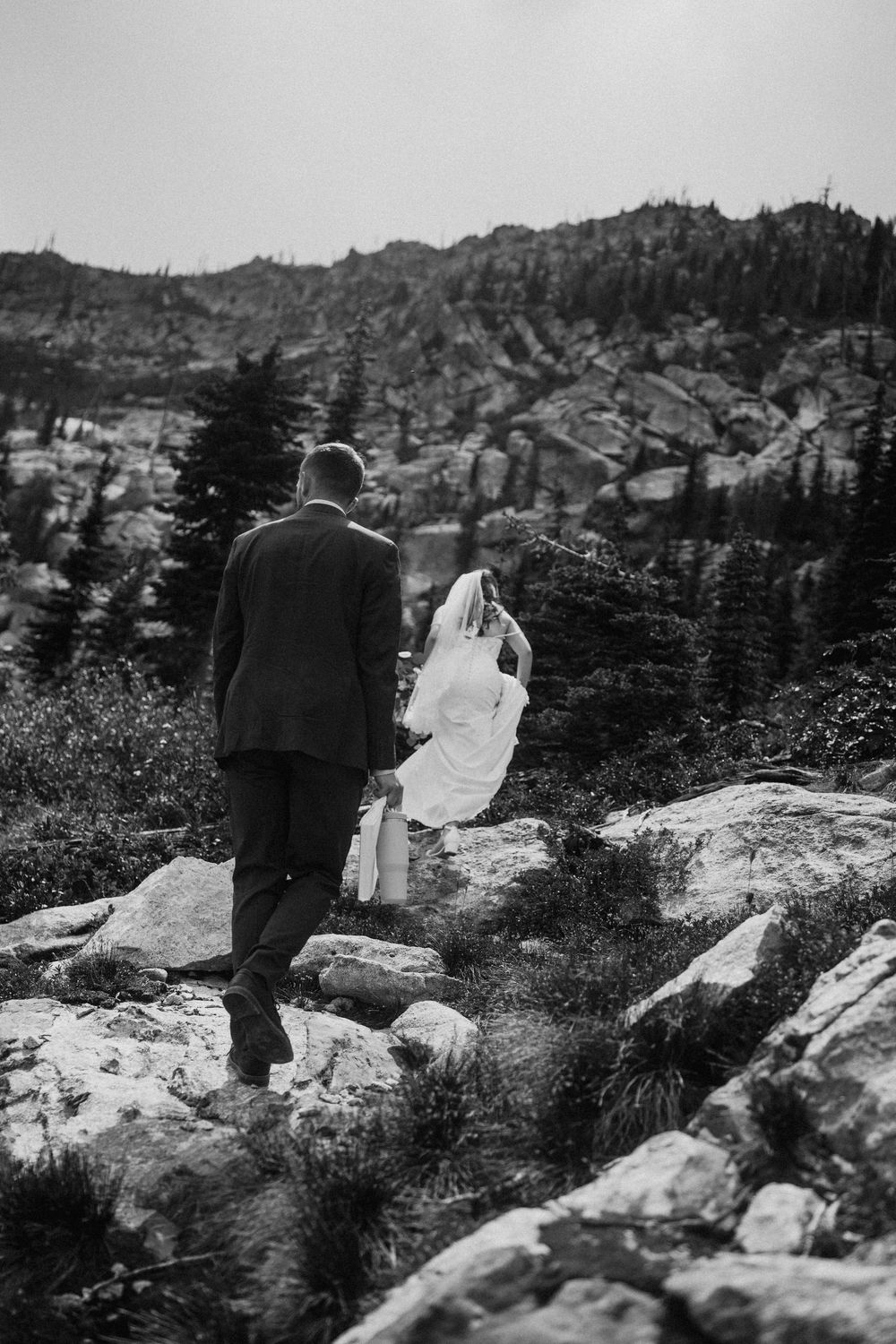 A couple in wedding attire walks along rocky mountain terrain in black and white.