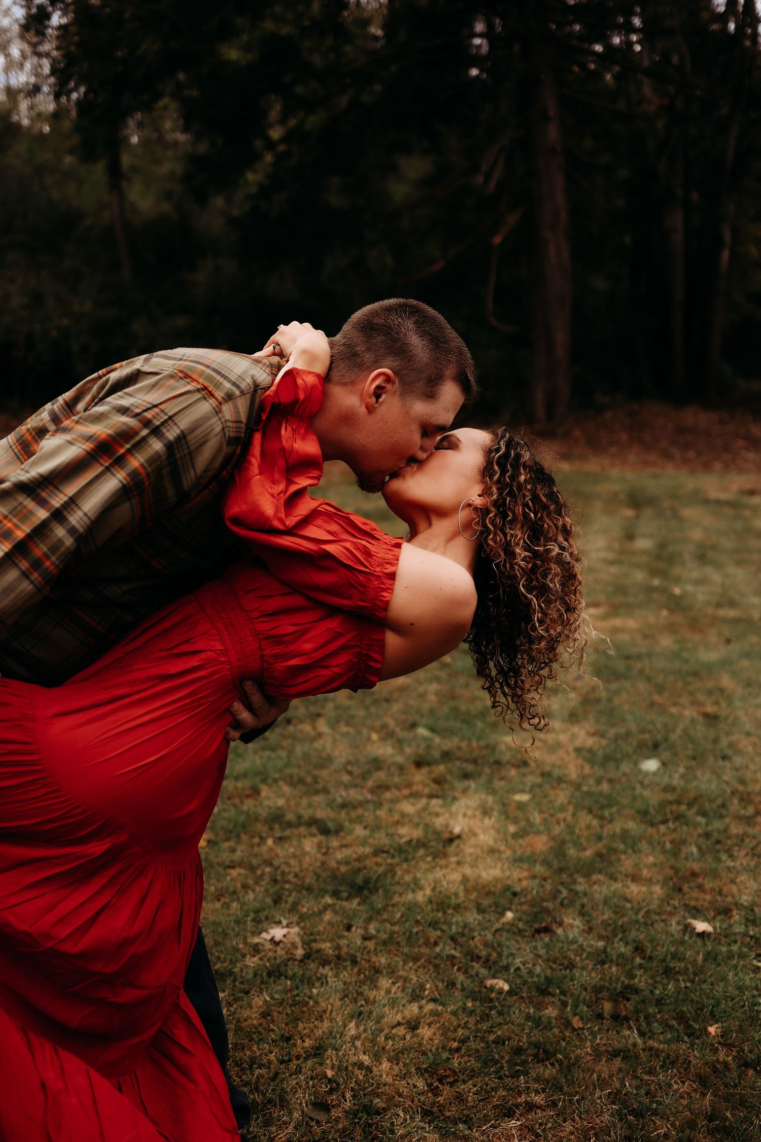 A romantic couple shares a passionate kiss in a wooded area, with the woman wearing a flowing red dress.