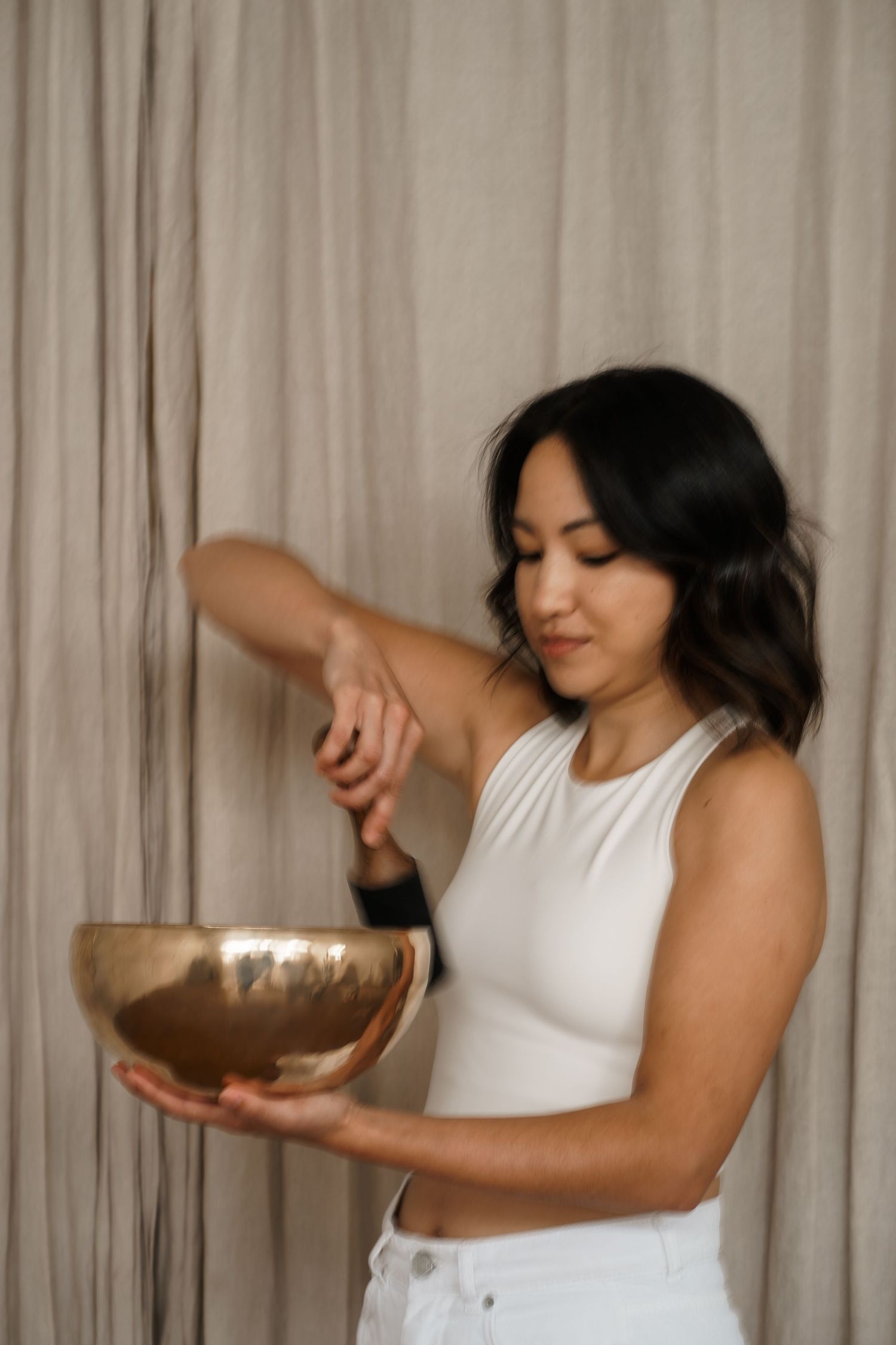 Person in white sleeveless top using a singing bowl for meditation practice against beige curtain background.