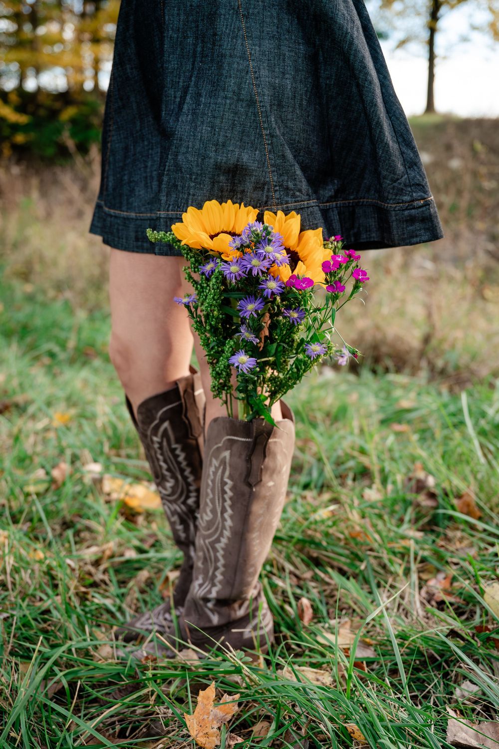 Brown cowboy boots with black dress holding wildflower bouquet of yellow and purple blooms in grassy field.