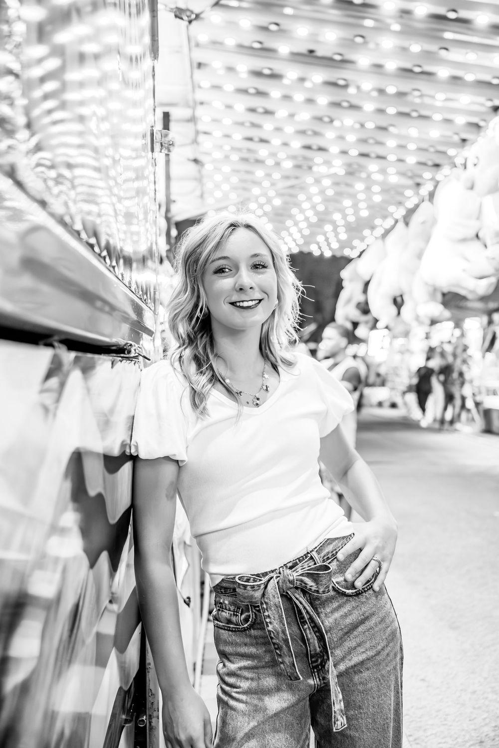 Black and white photo at a carnival with string lights overhead showing someone in a white top and jeans smiling.