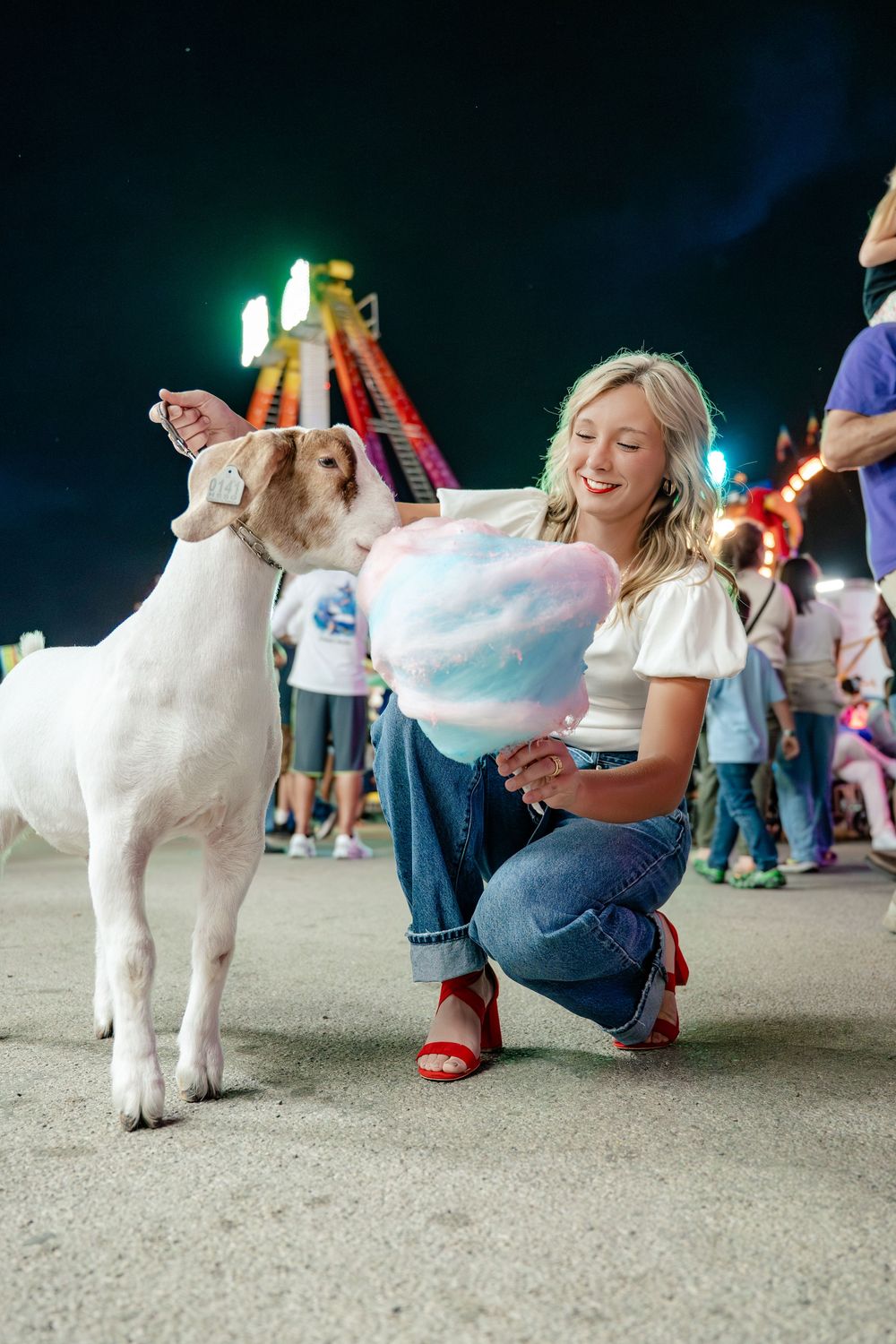 A person shares cotton candy with a small white goat at an outdoor event.