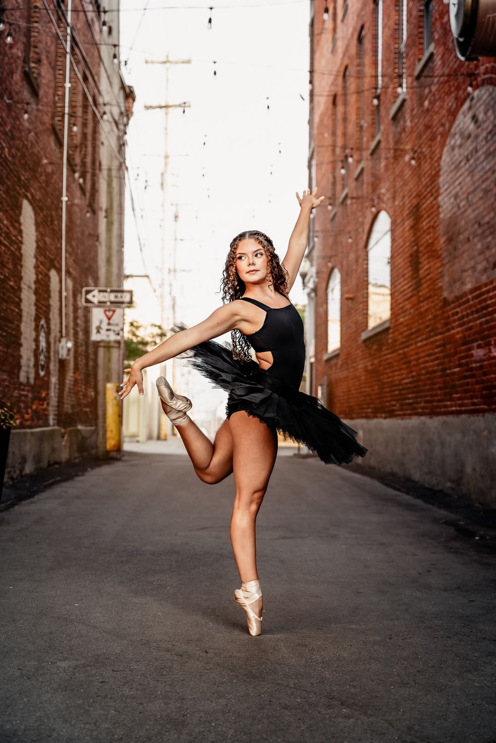 Ballet dancer performs graceful movements in black dress against urban brick alley backdrop.
