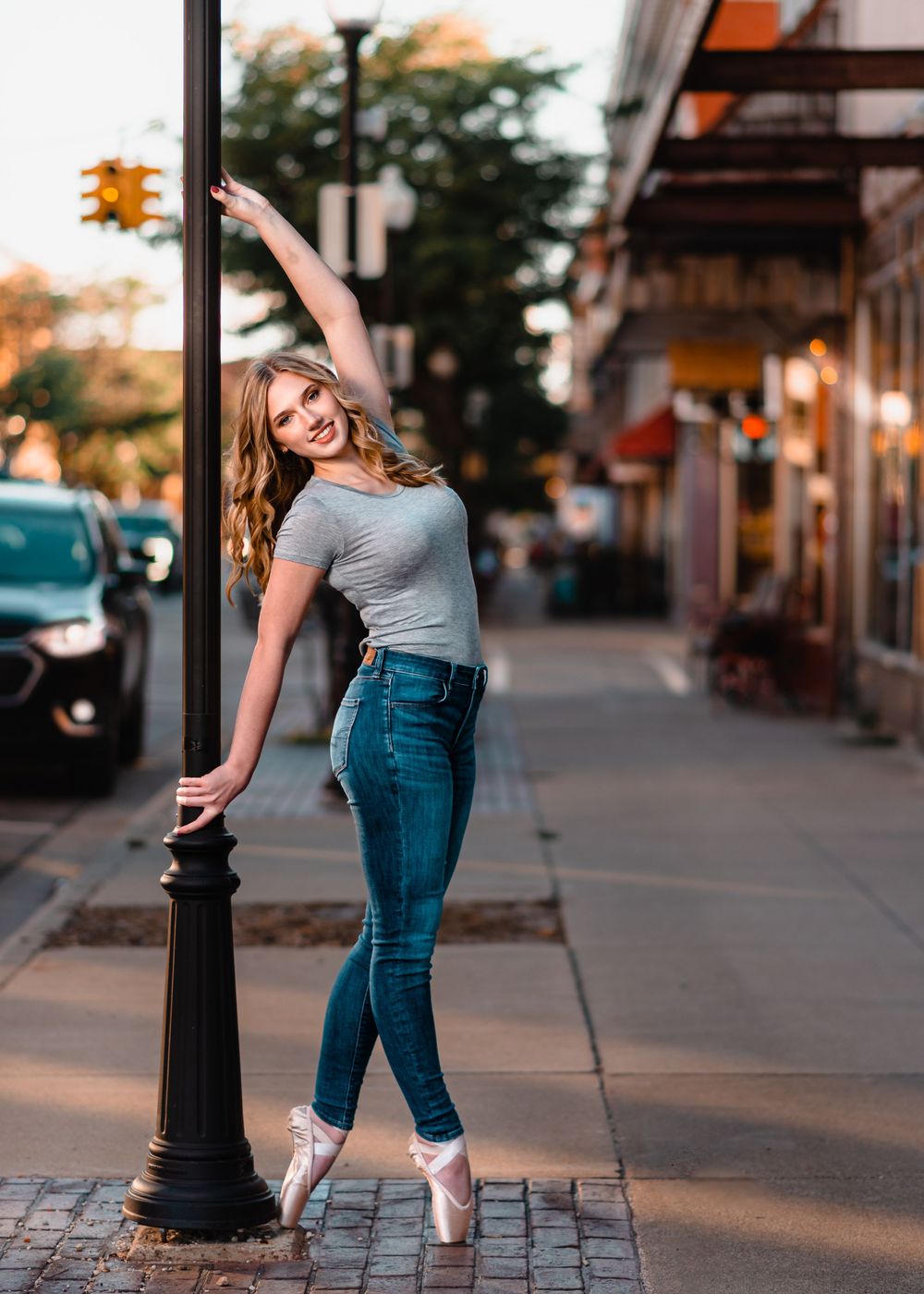 Ballet dancer in pointe shoes poses gracefully on city sidewalk at sunset wearing jeans and grey top.
