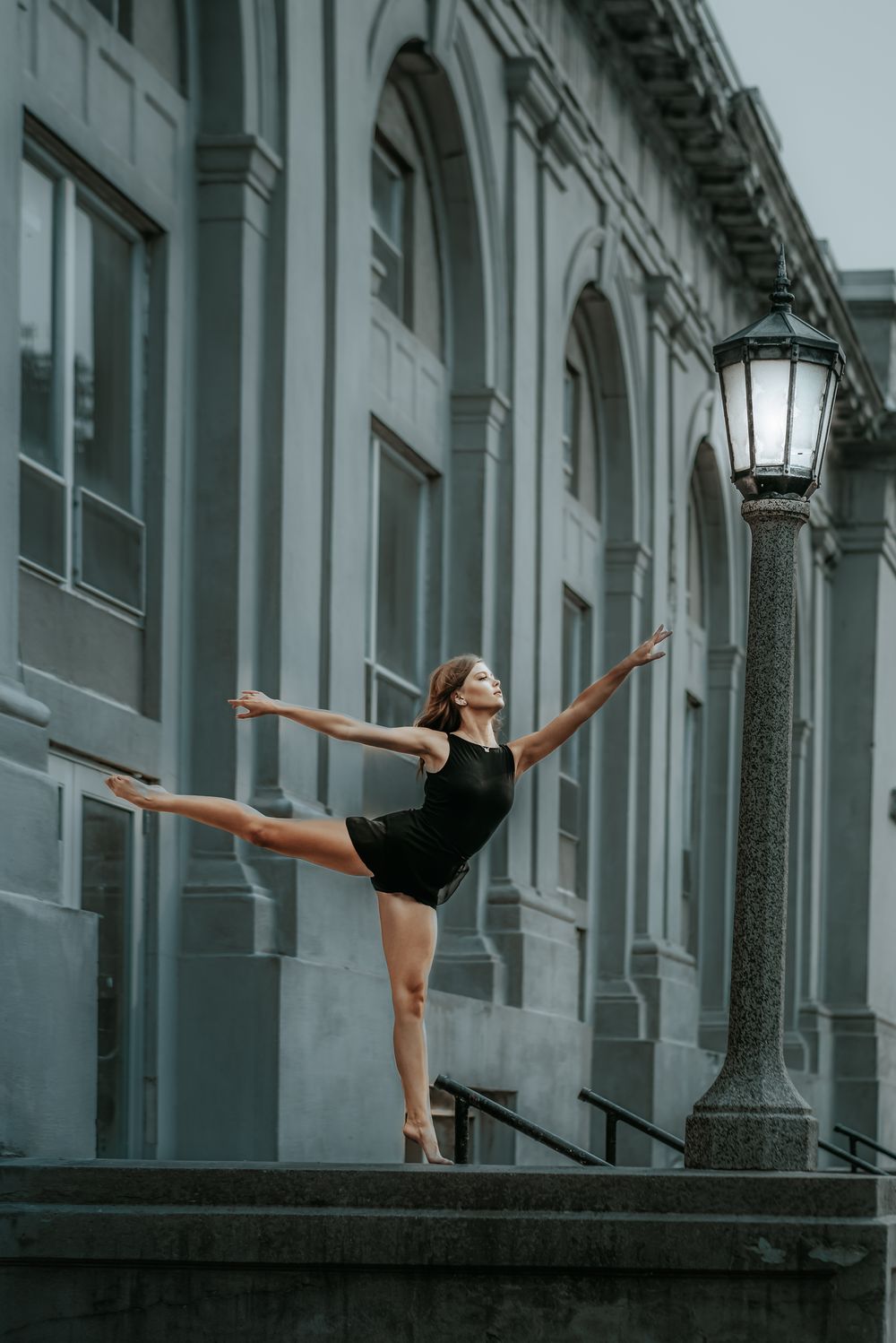 A dancer in a black leotard performs a graceful arabesque pose on stone steps outside a classical building with arches.