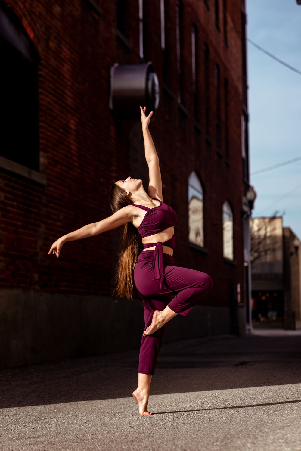 A dancer in burgundy athletic wear performs an elegant pose against a brick building backdrop in urban setting.