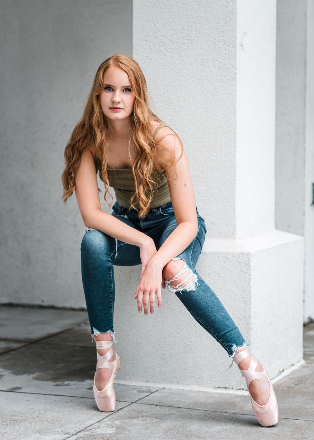 Ballet dancer in pointe shoes, distressed jeans and tank top poses against concrete wall in urban setting.