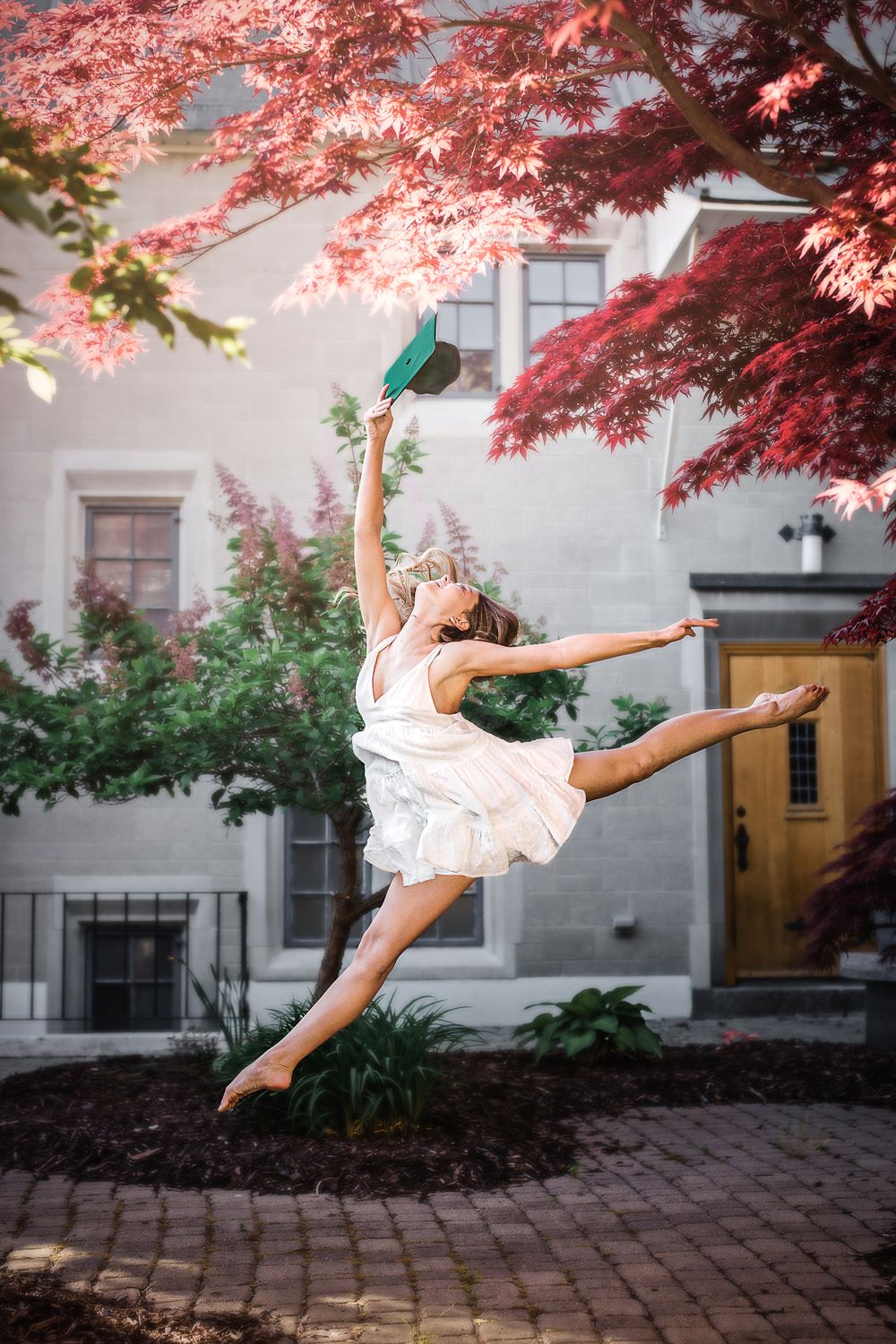 A dancer in white dress leaps gracefully beneath vibrant red Japanese maple trees against a stone building.
