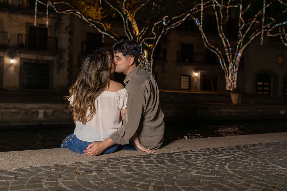 A couple embraces in a romantic moment on a cobblestone street at night with city buildings in the background.