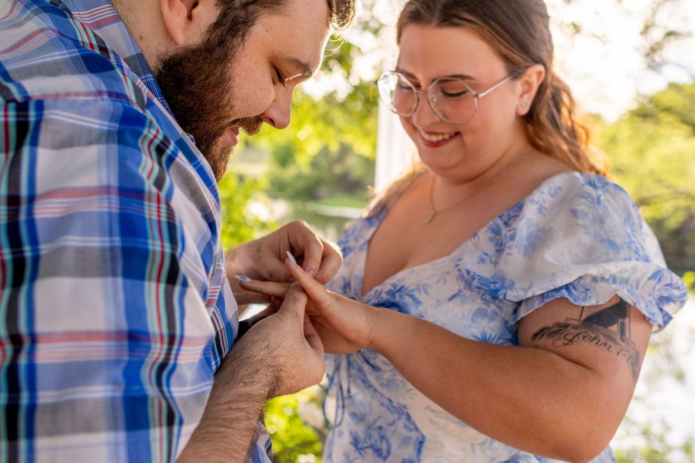 Couple sharing a romantic ring exchange moment outdoors, woman smiling wearing glasses and floral dress.