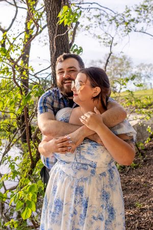 A bearded man in a plaid shirt hugs a smiling woman in a floral dress outdoors near trees.