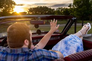 Couple relaxing in a classic convertible car at sunset, woman waving hand in the golden evening light.