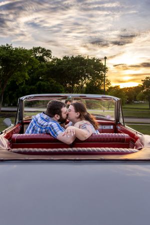 Couple kissing in the back seat of a vintage red convertible car at sunset with trees in the background.