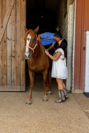Couple sharing a kiss beside a chestnut horse in a barn doorway, woman in white dress and cowboy boots.
