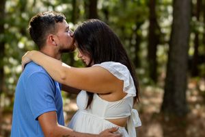 A couple embraces tenderly in a forest, man kissing woman's forehead while she rests her head on his chest.