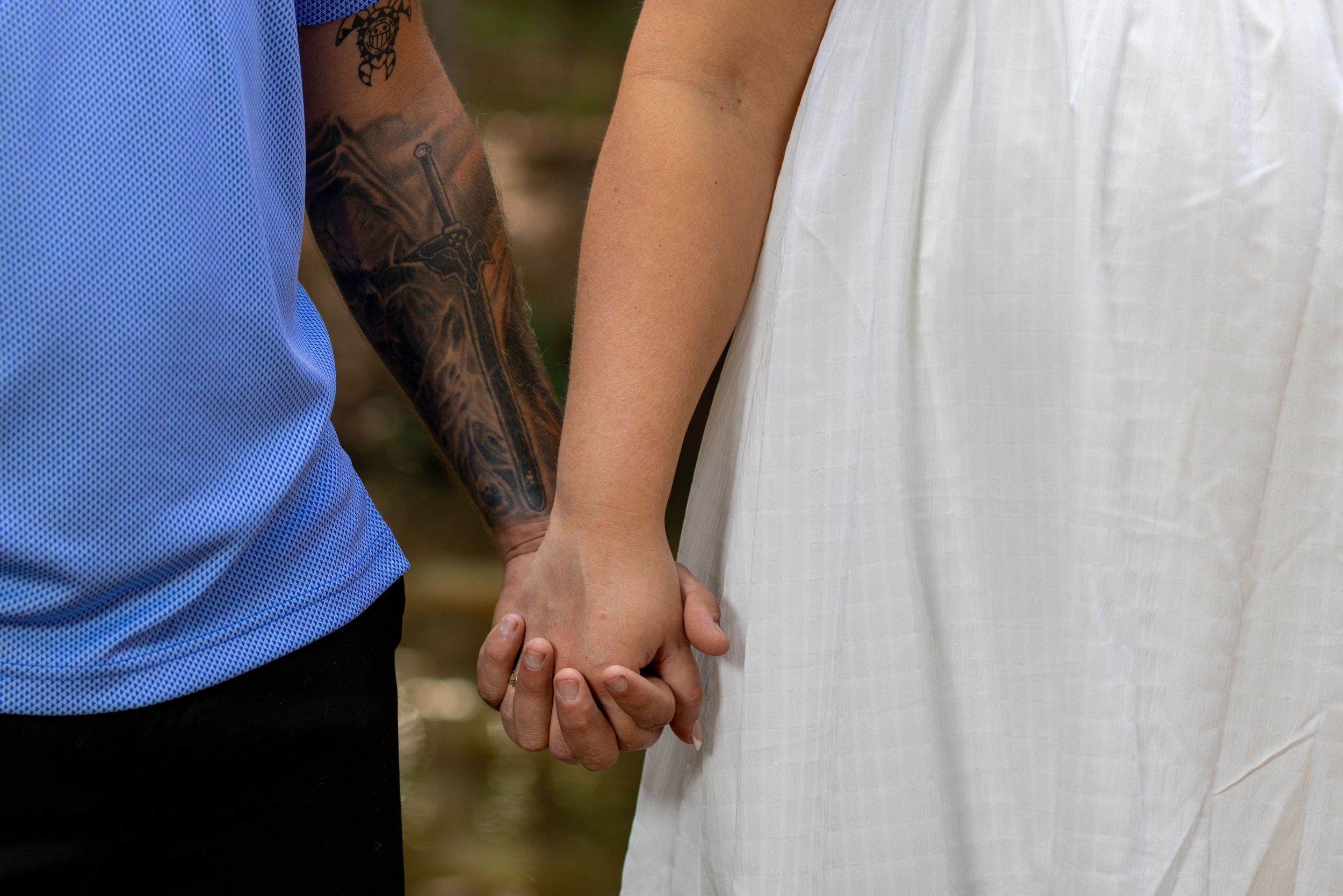 Couple holding hands, man with tattoed arm in blue shirt and woman in white dress, outdoor wedding photo.