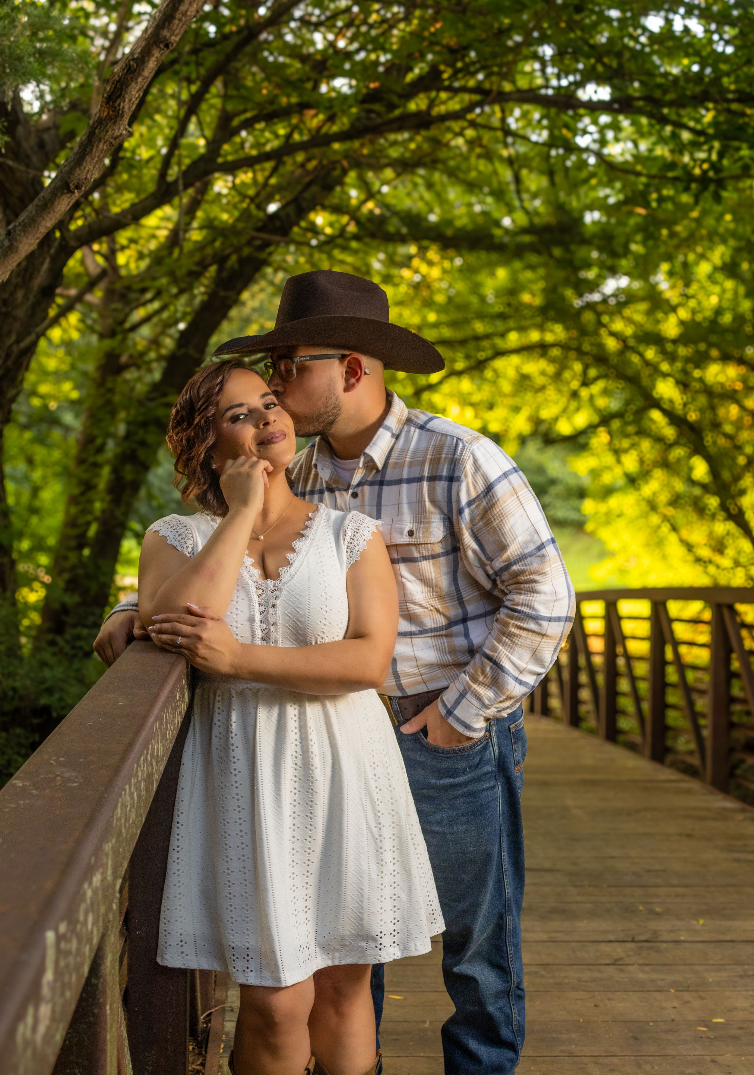 Couple sharing a romantic kiss on a wooden bridge surrounded by lush green trees, woman in white dress, man in cowboy hat.