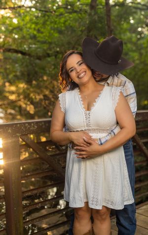Couple embracing on a wooden bridge at golden hour, woman in white lace dress smiling, man in cowboy hat kissing her cheek.