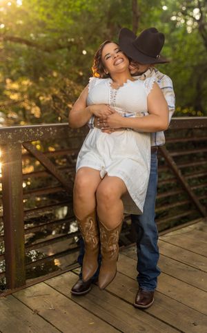 Couple embracing on wooden bridge, woman in white dress and cowboy boots, man in cowboy hat and jeans, surrounded by trees.