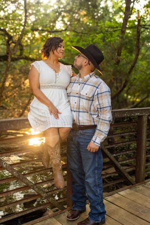 Couple in western attire sharing a romantic moment on a wooden bridge surrounded by lush green trees at sunset.