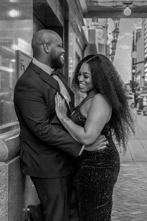 A couple in formal attire share a romantic moment on a city street, laughing and embracing in black and white.