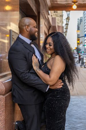A couple in elegant evening wear embraces while posing against a brick wall in an urban setting.