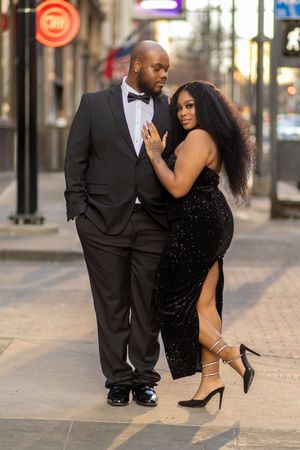 A romantic couple in black formal wear embraces on an urban sidewalk during evening hours.