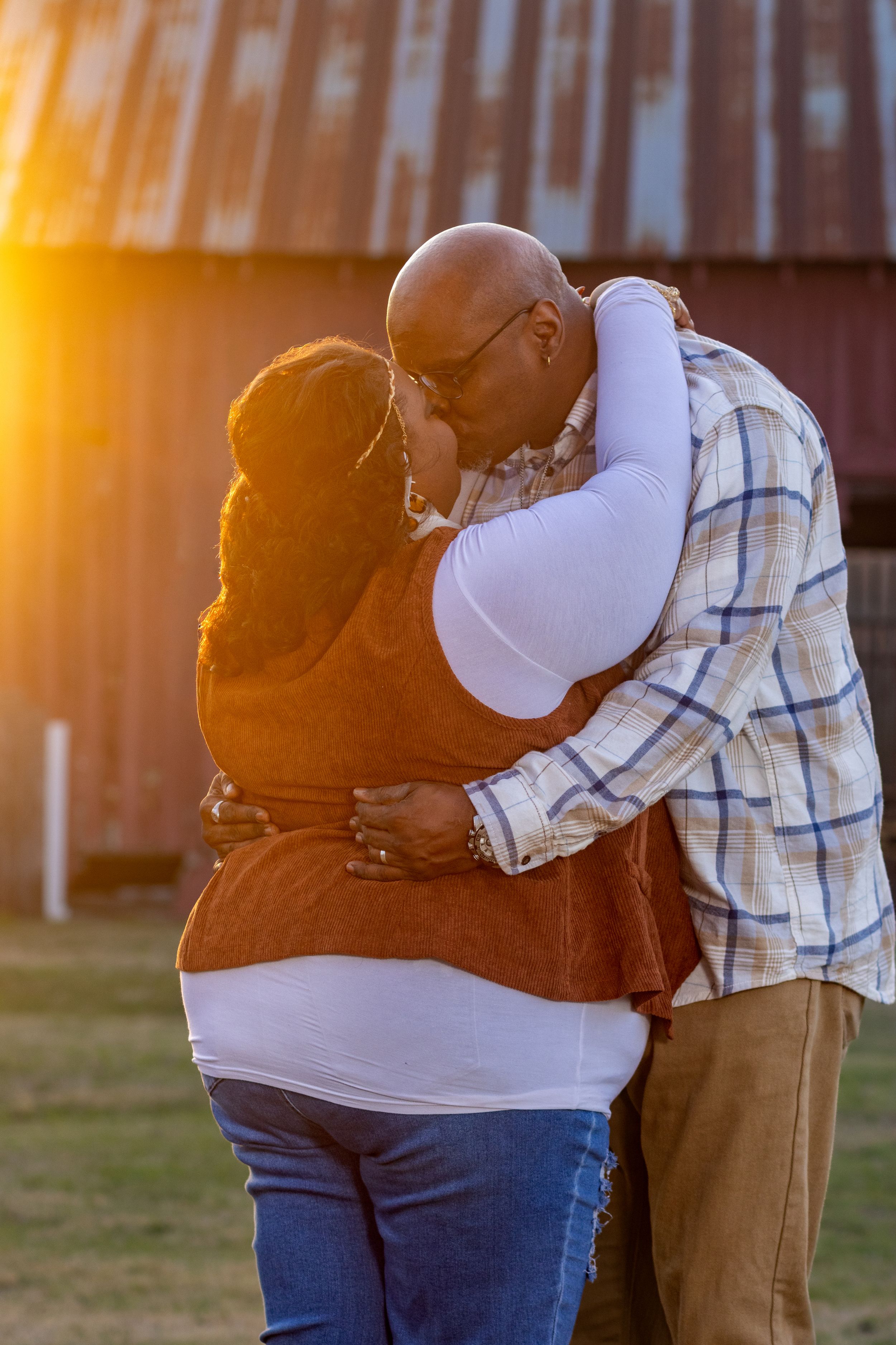 A couple embraces in warm sunset light while standing on grass, creating a romantic silhouette against the golden sky.