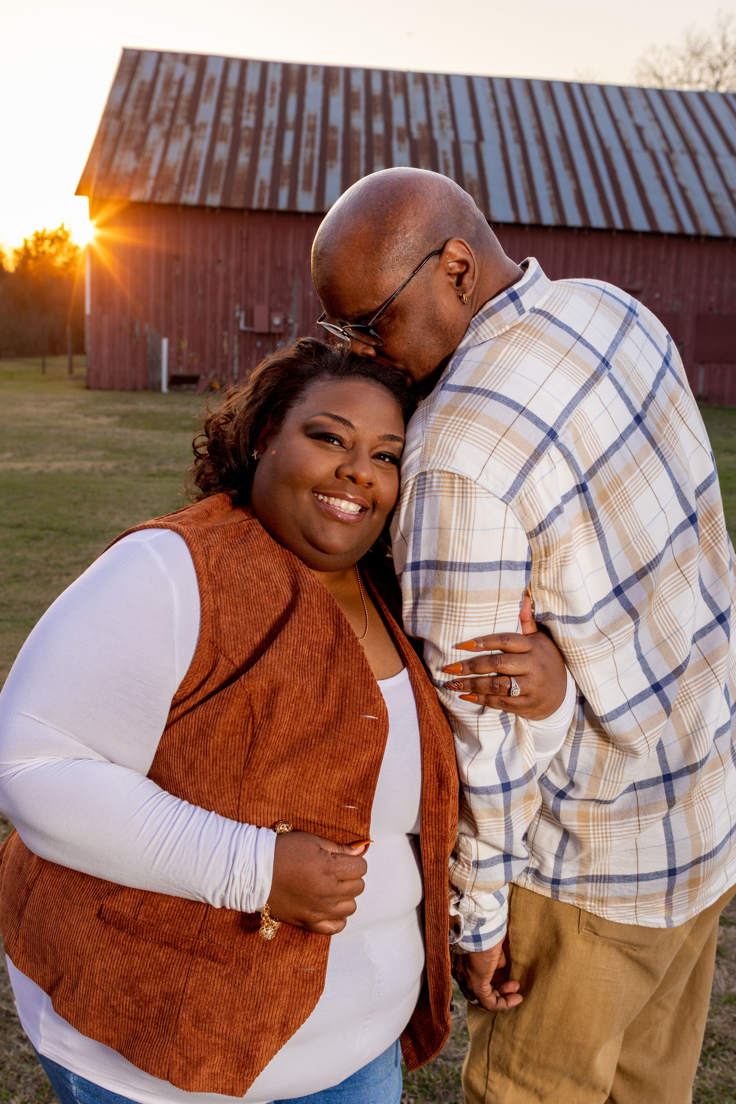 Romantic couple embraces at sunset in front of a rustic barn wearing coordinated brown and white outfits.