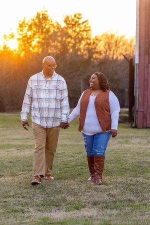 Couple walking hand in hand through a grassy field at sunset with warm golden light behind them.