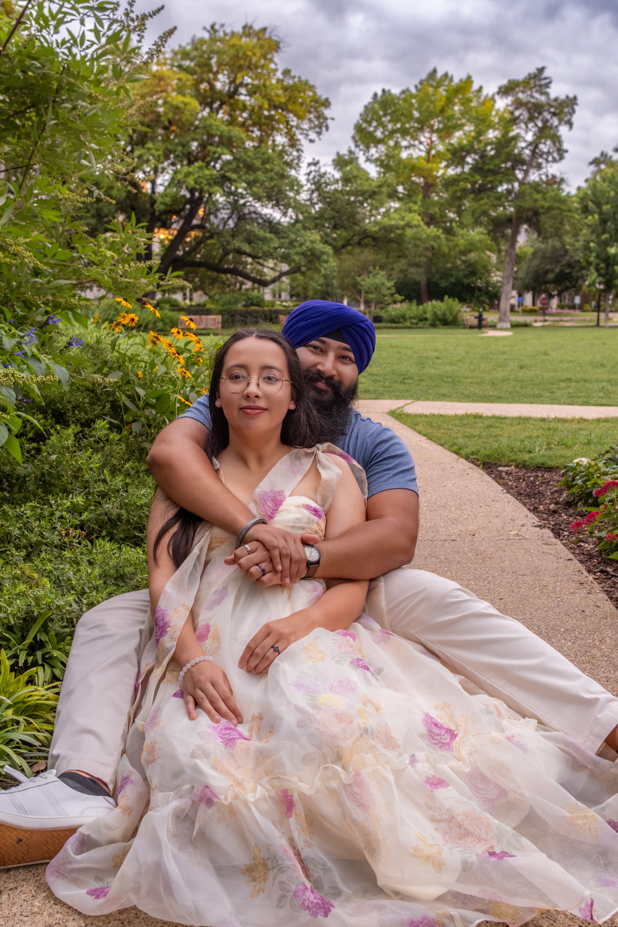 A couple sits together in a lush park, woman in floral saree, man in blue turban embracing her warmly.