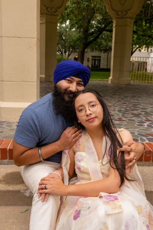 A Sikh man in a blue turban embraces a young woman in a floral dress, posing together under an archway.