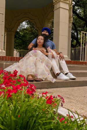 Sikh couple sitting on steps near floral garden, man in blue turban embracing woman in floral gown.