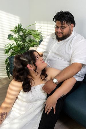 A couple shares a tender moment indoors, woman in white dress looks up at bearded man in white shirt near a plant.