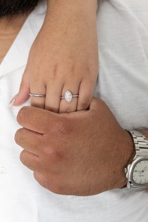 Close-up of a woman's hand wearing a marquise diamond halo engagement ring, resting on a man's hand with a silver watch.