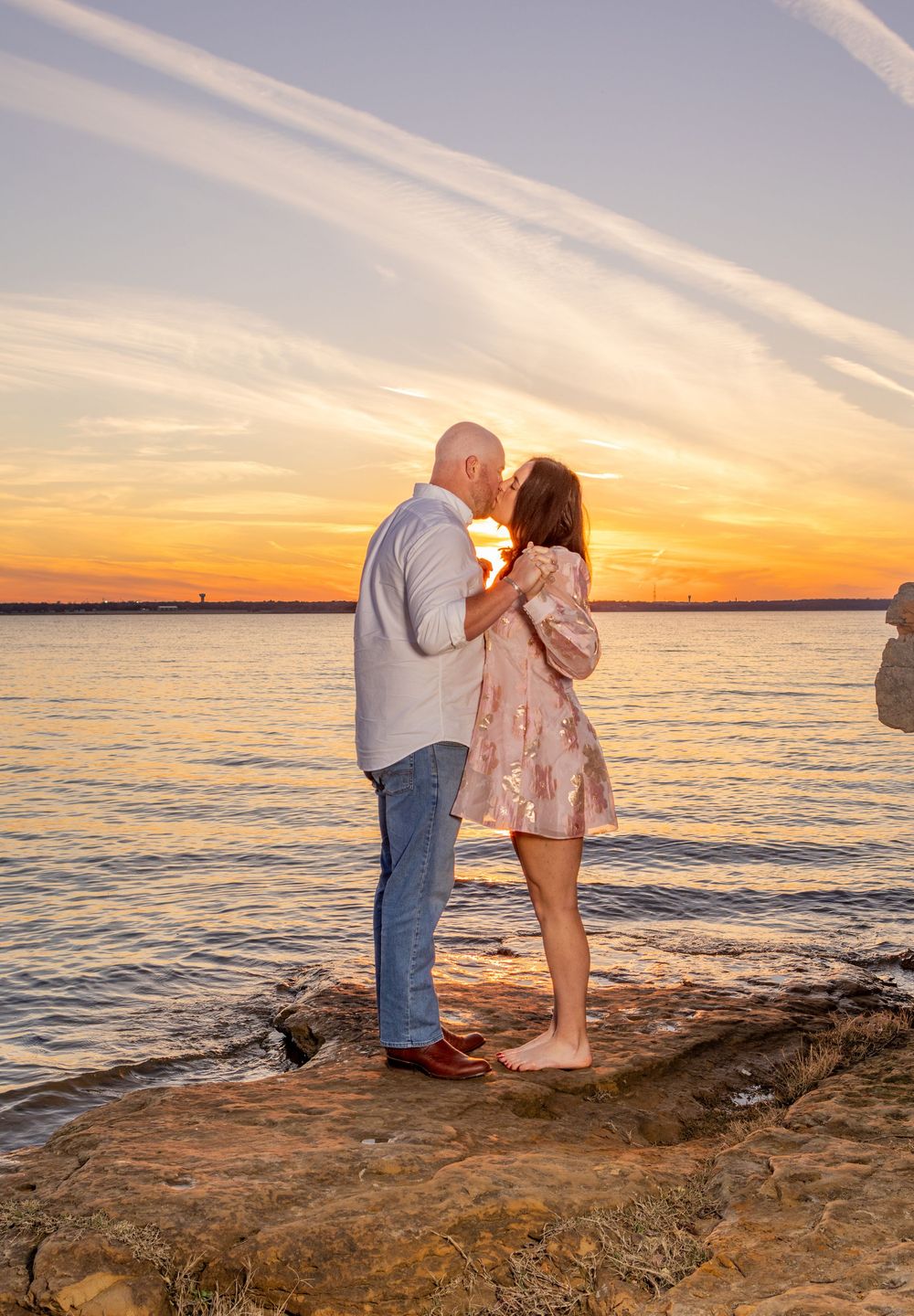 A romantic couple shares a tender moment on rocky shoreline during golden sunset over calm waters.