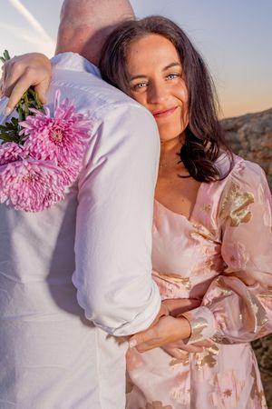 Romantic couple in pink and white outfits embrace while holding a pink chrysanthemum flower during sunset.