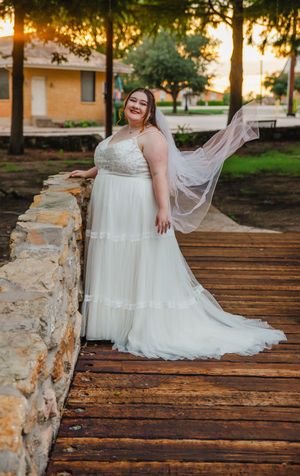 A bride in a white gown sits on rustic wooden planks beside a stone wall at sunset.