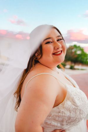 A bride in a white lace dress poses joyfully against a sunset sky.