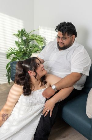Pregnant couple in white outfits sharing a tender moment on a teal couch with a green plant in the background.