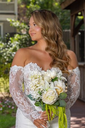 Bride in off-shoulder lace gown holding white floral bouquet with green ribbons, posing outdoors in natural light.