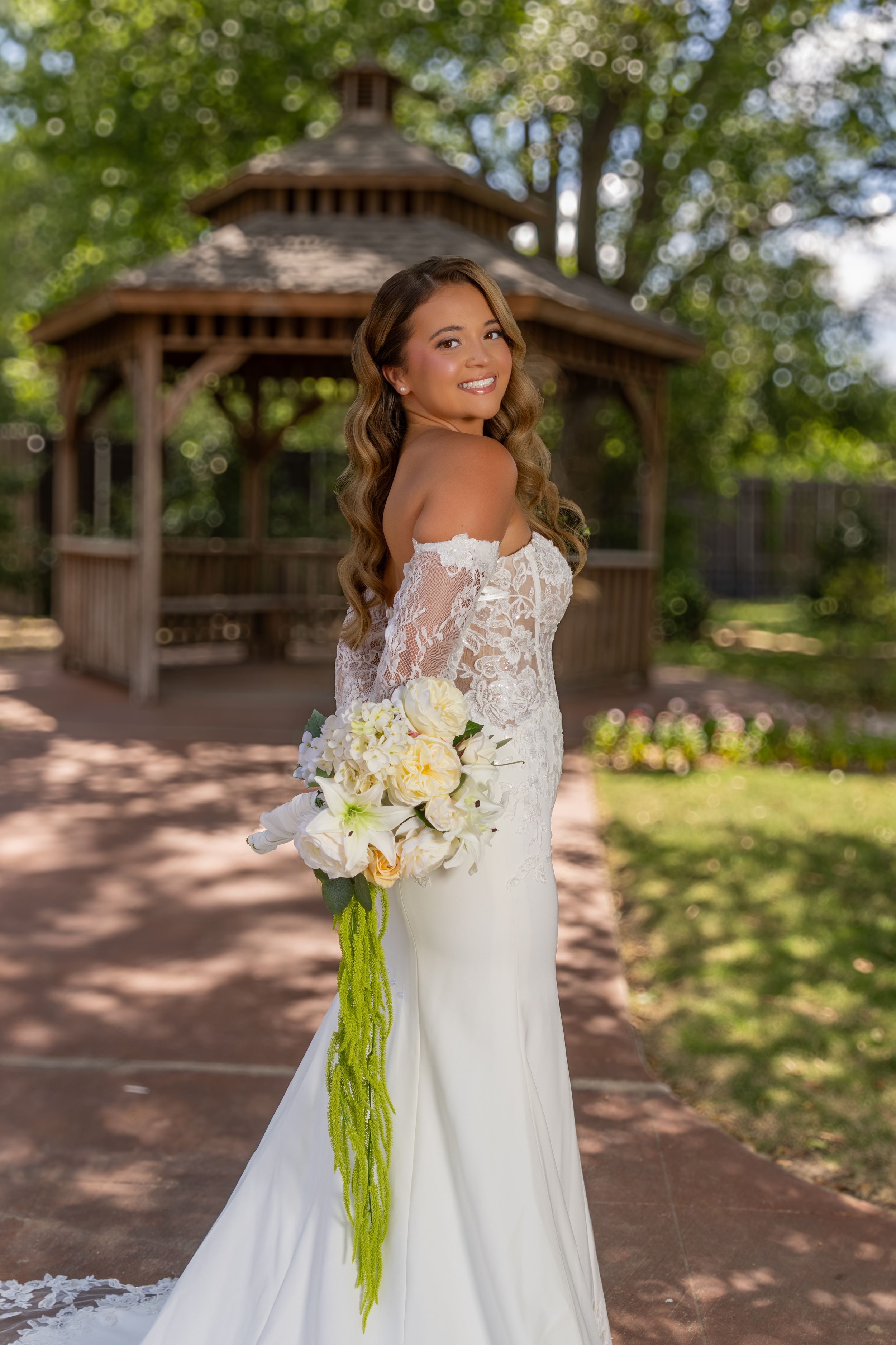 Smiling bride in lace long-sleeve wedding gown holds white floral bouquet outdoors near a wooden gazebo.