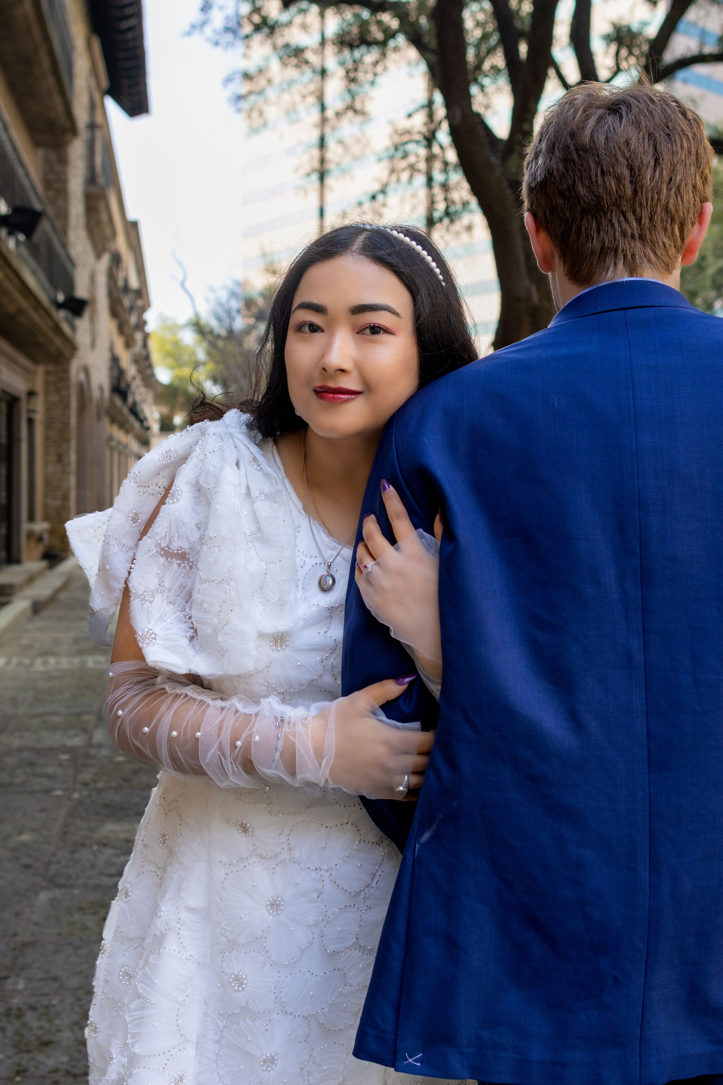 A couple in formal attire, with a white lace dress and navy suit, pose together on a stone pathway.