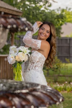 Smiling bride in off-shoulder lace wedding gown holding white bouquet in garden setting.