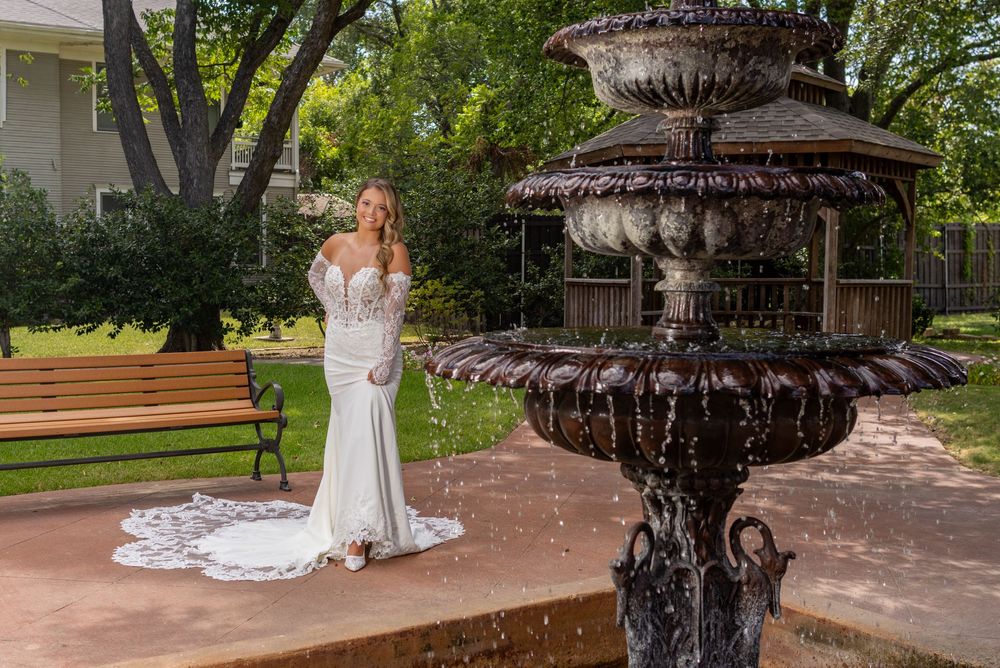 Bride in white lace gown poses near ornate tiered fountain in lush garden setting.