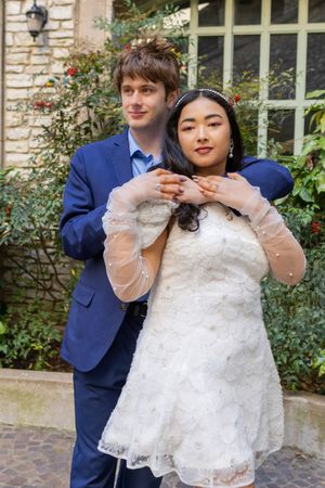 A newlywed couple poses together outdoors, with the bride wearing a white lace dress and groom in a navy blue suit.