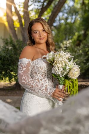 Bride in off-shoulder lace wedding gown holding white floral bouquet, posing outdoors with golden sunlight filtering through trees.